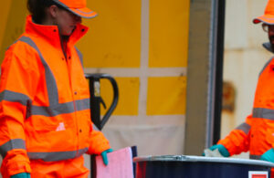 Two individuals transporting a barrel, both dressed in high-visibility clothing.